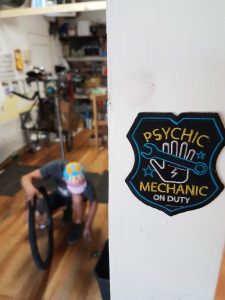 A mechanic bent over a bike wheel in the fuzzy back ground, a patch statin "Psychic mechanic at work' focused in the foreground